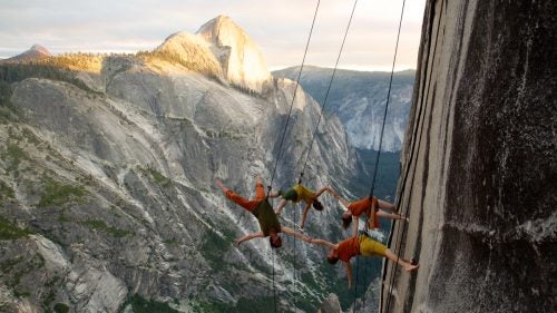 In 1989, choreographer Amelia Rudolph was clinging to a sheer granite wall in California’s Sierra Nevada when she wondered what it would look like to dance there. Soon after, Rudolph founded Bandaloop, a dance group that has performed on numerous rock walls, as well as Seattle’s Space Needle, skyscrapers in South Korea, and dozens of other sideways stages, sometimes for public audiences numbering in the tens of thousands. Beginning this June 16, six dancers and a nine-person support crew headed into Yosemite’s wilderness for ten days of hiking and pirouetting. The pinnacle of the journey was a performance on the face of 8,500-foot Mount Watkins, a stage that offers a rare perspective of Half Dome and an audience of birds, bugs, squirrels, and perhaps a bear or two.
Photo: Bandaloop dancers on Mount Watkins, nearly 3,000 feet above the valley floor. Rudolph’s goal with the group has been to challenge preconceptions about modern dance. The performers make it look easy to fly off the wall, but their abdominal muscles are often screaming as they fight gravity to hold their bodies perpendicular to the face.