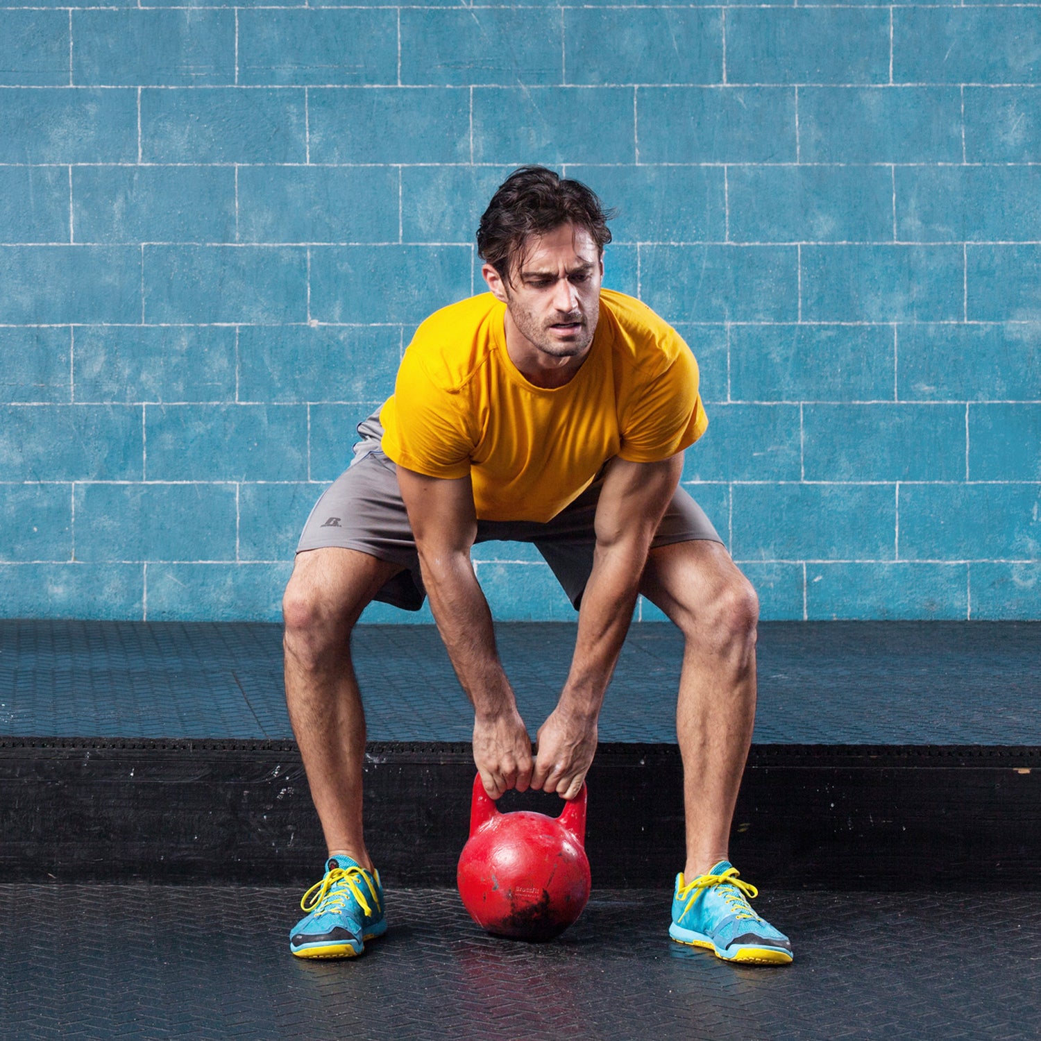 Male athlete strains at the beginning of a lift during a crossfit workout with a kettle bell. The background is blue brick and there are several used kettle bells behind him