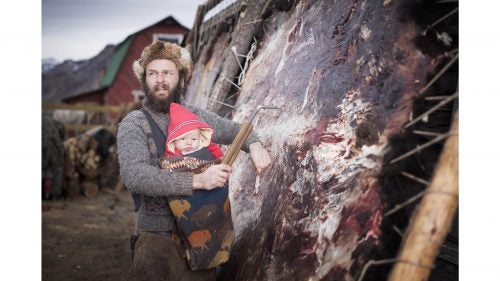Alex carries his son as he works at fleshing a hide for tanning.