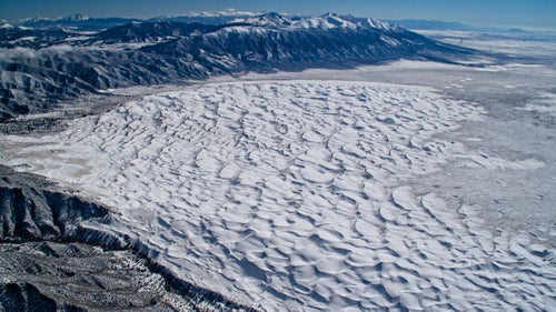 For those who visit Great Sand Dunes National Park in the summer tourist season with sand temperatures approaching 140 degrees, it’s hard to believe the place ever gets cold. The San Luis Valley of Colorado can indeed be one of the coldest areas of the country in winter, trapped in powerful inversions of cold air.
On this particular February afternoon, the dunes were covered in a layer of snow as I wore double layers of ski gear, sticking the camera out the window in temperatures of minus 20 degrees in pursuit of fourteeners on the horizon.
From the book “Above the Summit: An Antique Airplane Conquers Colorado’s Fourteeners.”
