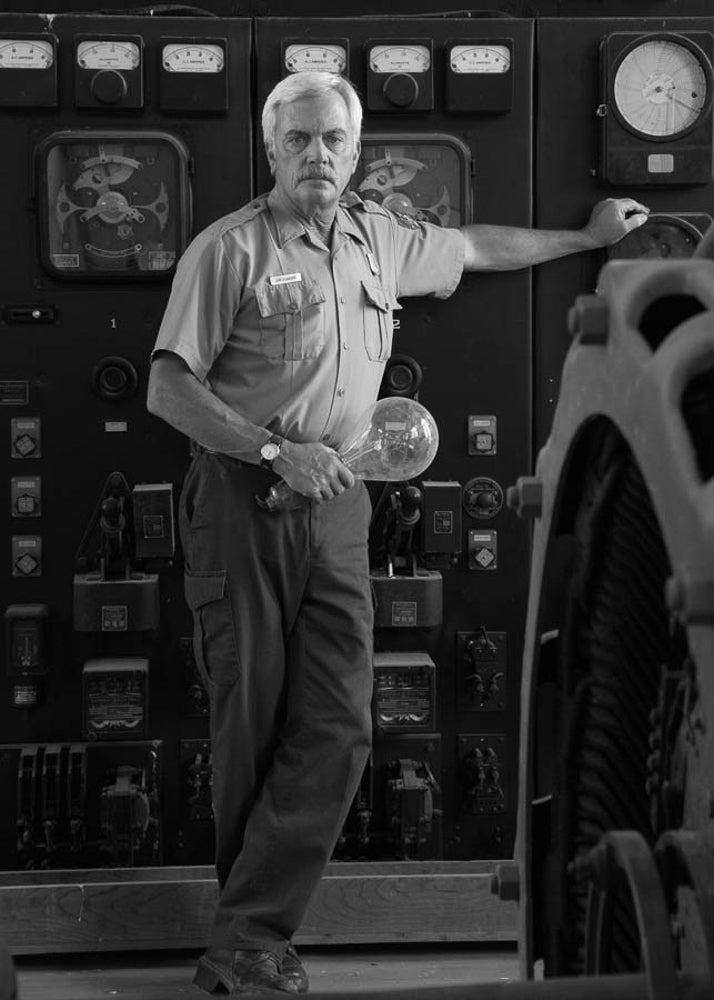 Jim stands in front of a generator instrument panel. Formerly used in Yellowstone National Park, the generator was powered by the energy of falling water through domestic water piping to supplement electricity production for the local utility company.