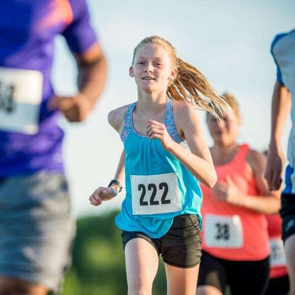 A large group of people are running a race outdoors. They are wearing athletic clothing and shoes, and they have racing numbers. They are running towards the camera. A Caucasian girl is in focus.