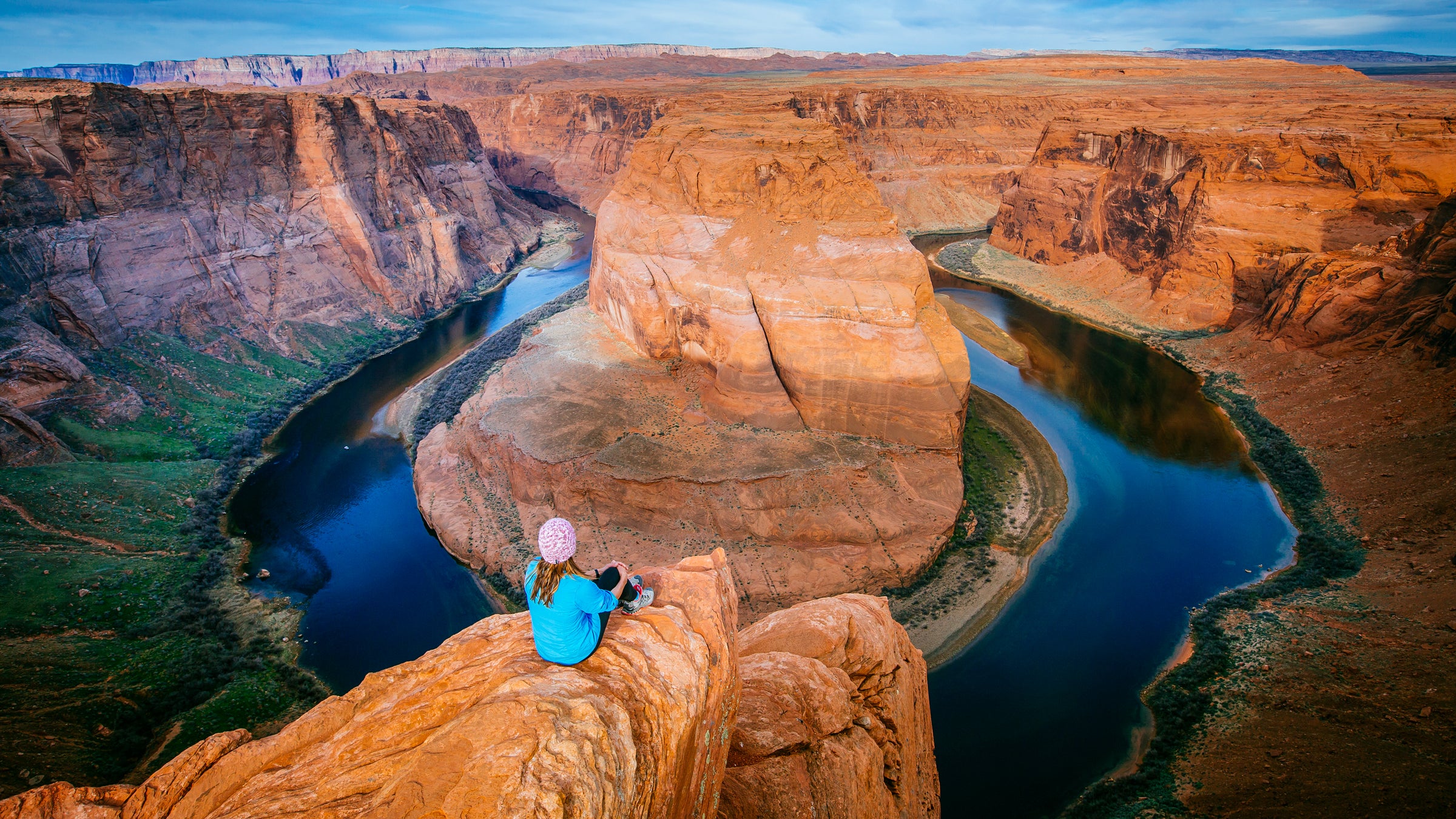 A woman sits on a cliff edge above the Horseshoe Bend of the Colorado River.