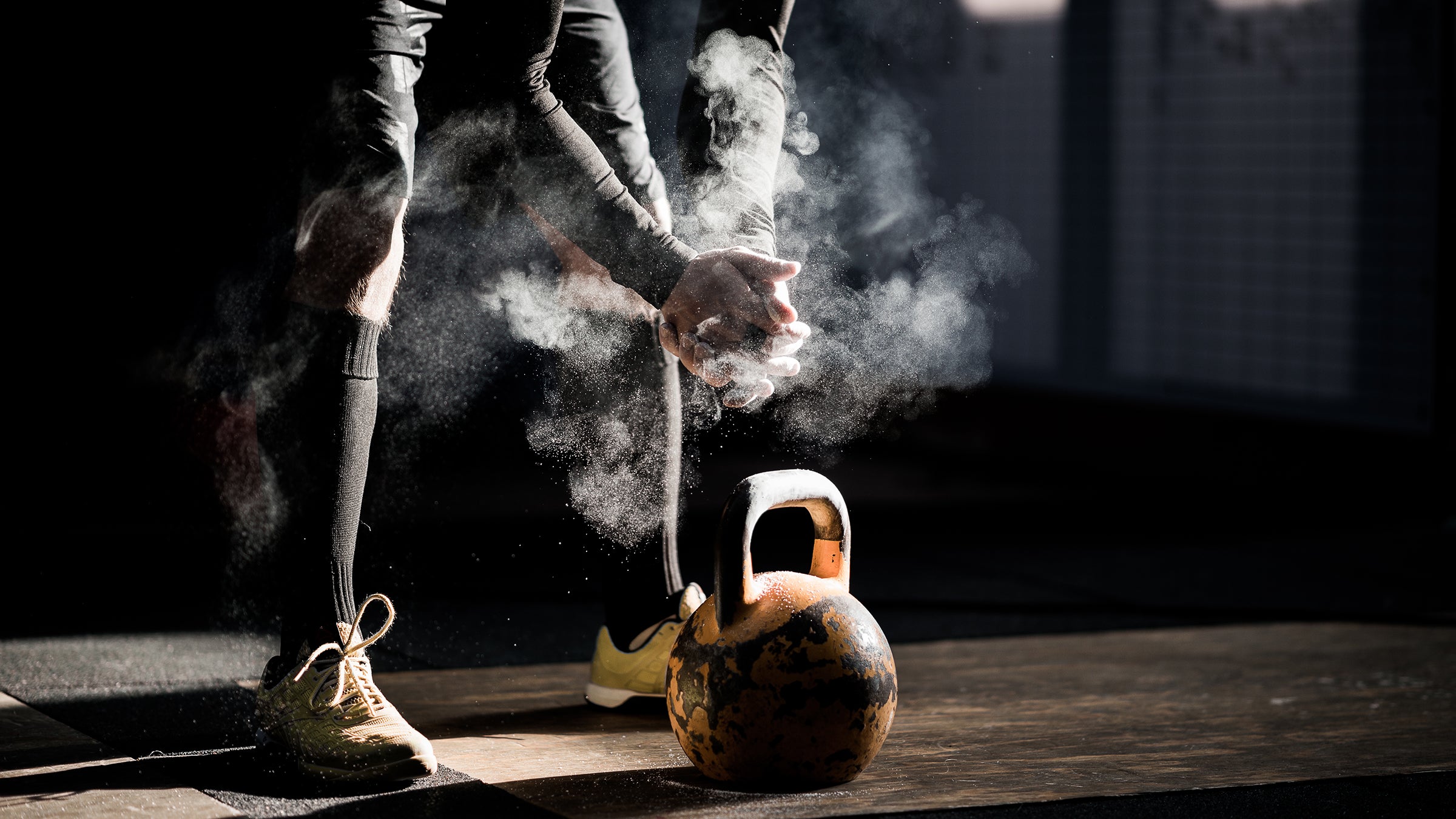 A person in a dimly lit gym chalks up their hands before lifting a kettlebell to complete a kettlebell workout plan.