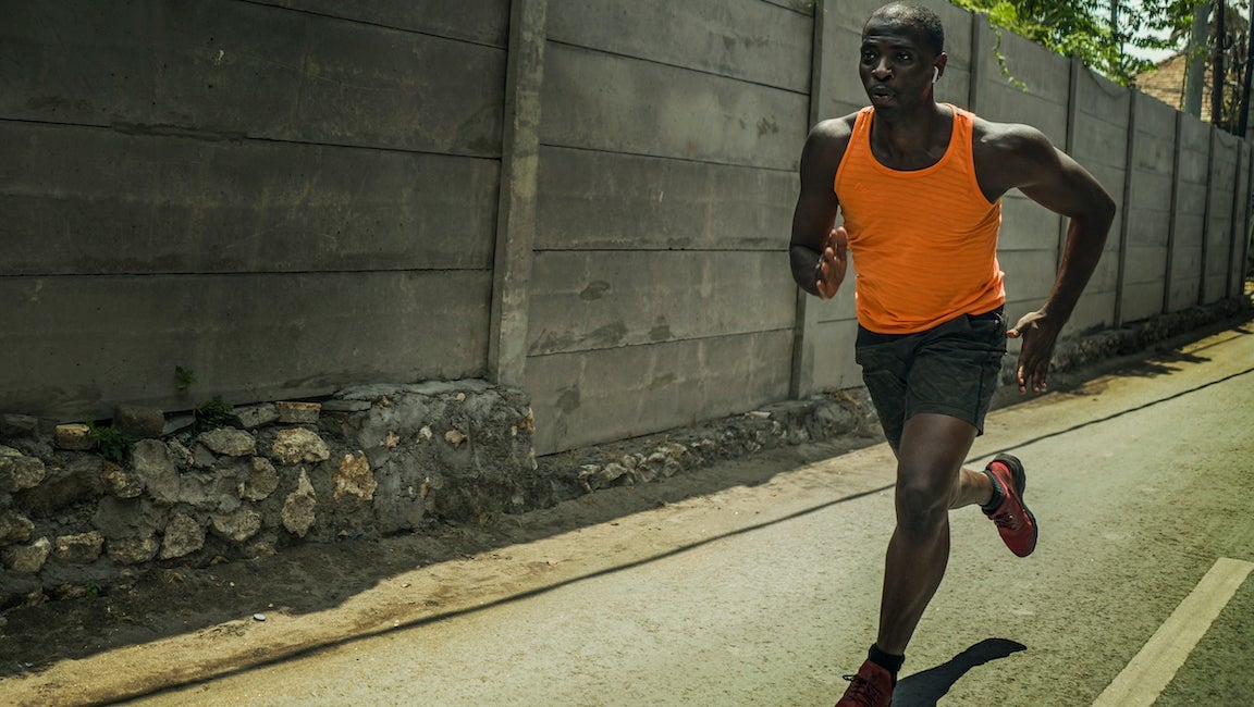 Fit professional sport man with athletic body running on city street pushing hard under harsh sun in urban background.