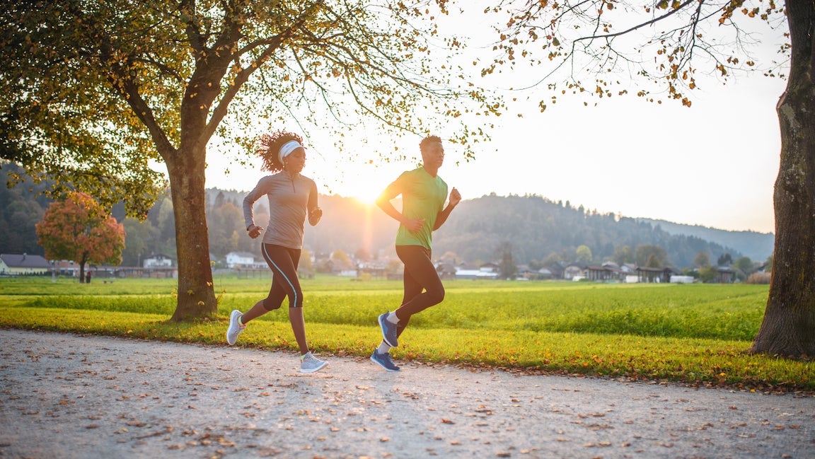 Full length view with lens flare of male and female sportspeople in 20s and 30s running past camera on pedestrian path with autumn leaves on the ground.