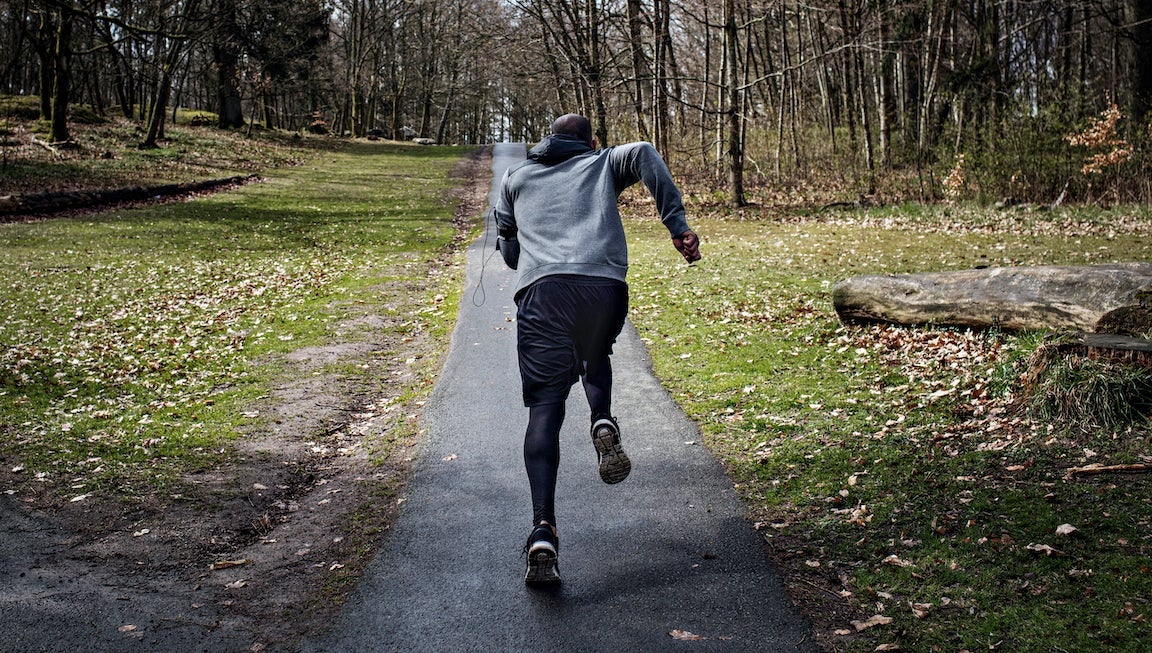 Full length rear view of determined male athlete jogging on narrow street in forest.