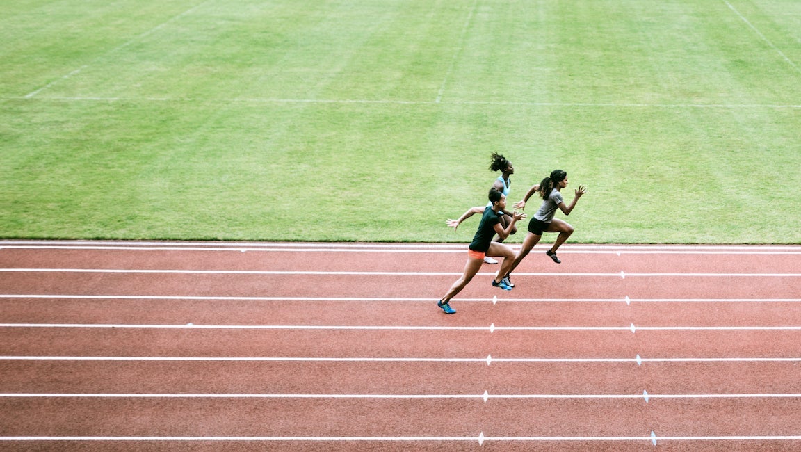 Young adult women sprint down the track, viewed from high in the stadium seating. Wide high angle view. Training for track and field competition.