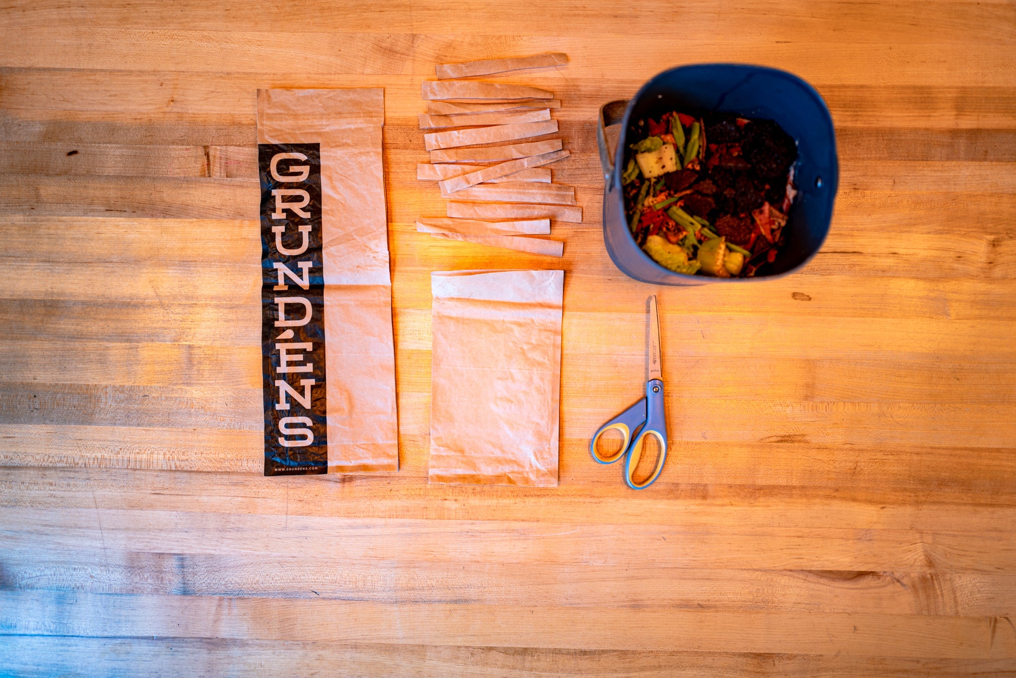 Grundens compostable packaging: A brown bag, partially cut into strips lays on a wooden surface next to a bucket of vegetable scraps and a pair of scissors