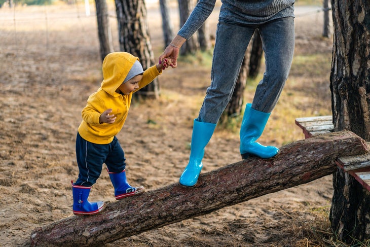 active-baby-boy-3036405 Toddler boy in yellow sweatshirt and royal blue rainboots holds the hand of an adult in gray with turquoise rain boots as they walk up a log to a…