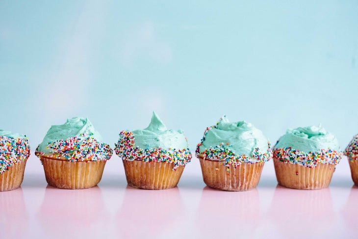 brooke-lark-pGM4sjt_BdQ-unsplash Light blue frosted vanilla cupcakes with colored ball sprinkles on the edge sit on a pink and blue background.