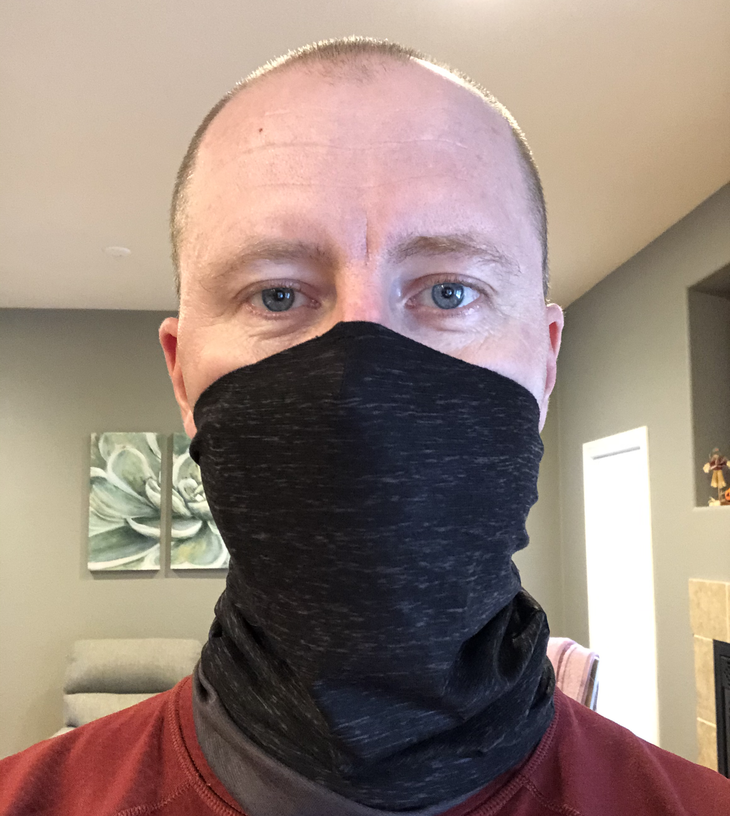 "Man staring into camera wearing black face mask tube and red shirt; furniture and wall hangings behind him."