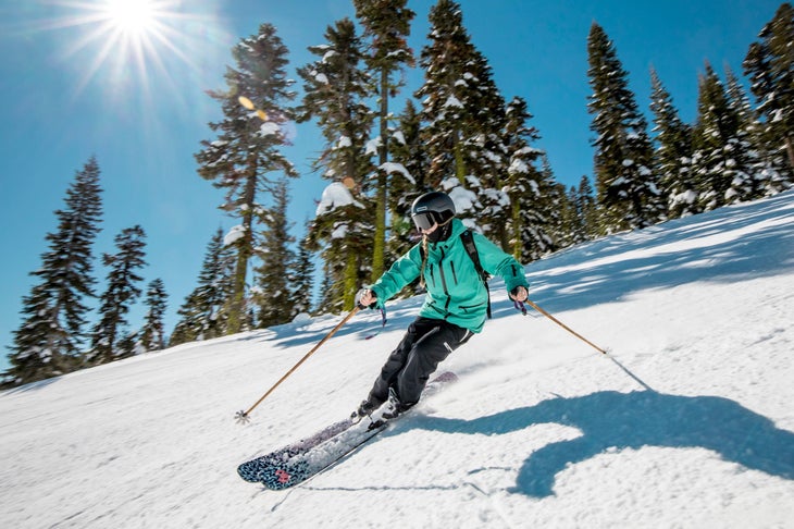DG4A2561 Woman in blue jacket and black snowpants skis on Coalition Snow skis with sun and frosted trees in the background.