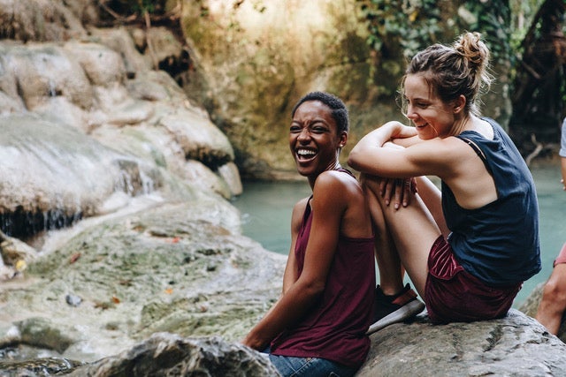 diversity4 Two smiling women sitting on a rock in front of a pool of water. One woman is Black with short hair and a dark red tank top, the other is white with…
