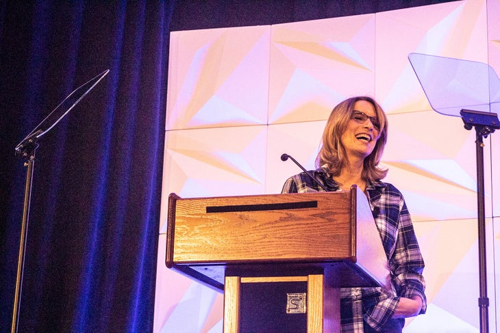 emily newman camber breakfast woman with long blonde hair wearing glasses and white t-shirt with flannel shirt stands in front of a podium giving a speech.