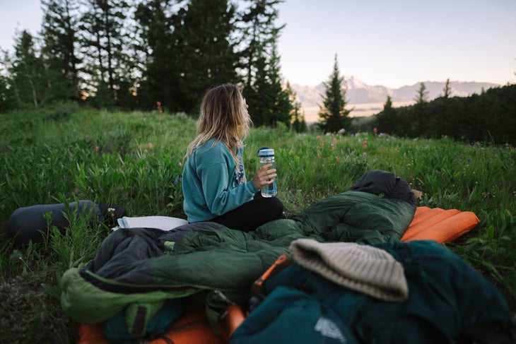 HP_Recon_5712 Woman sitting on sleeping bag in green field, looking into the distance. Mountains in the background.