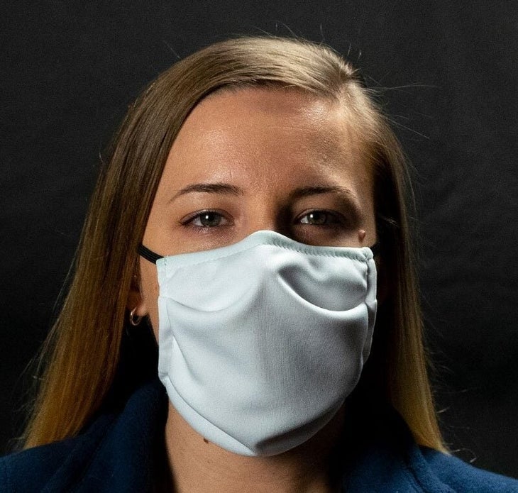 "Woman with long brown hair wearing white face mask; black wall behind her"