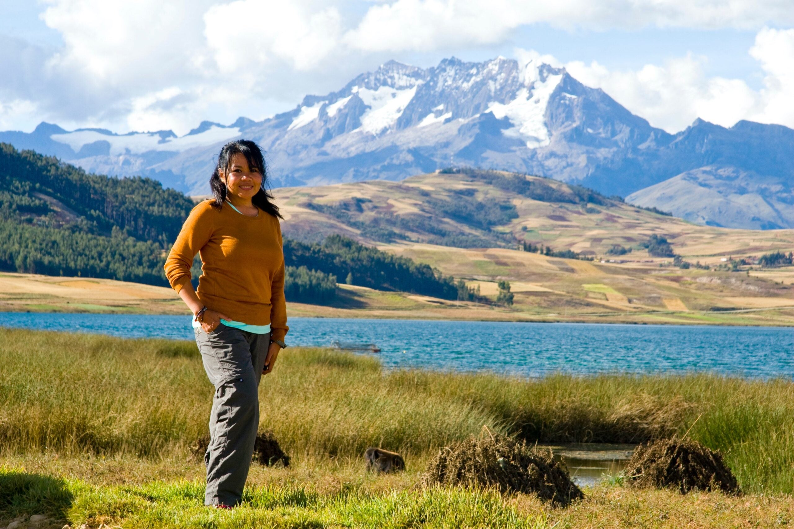 woman in gray hiking pants and orange sweater standing in the grass in front of a lake with snowy mountains in the distance