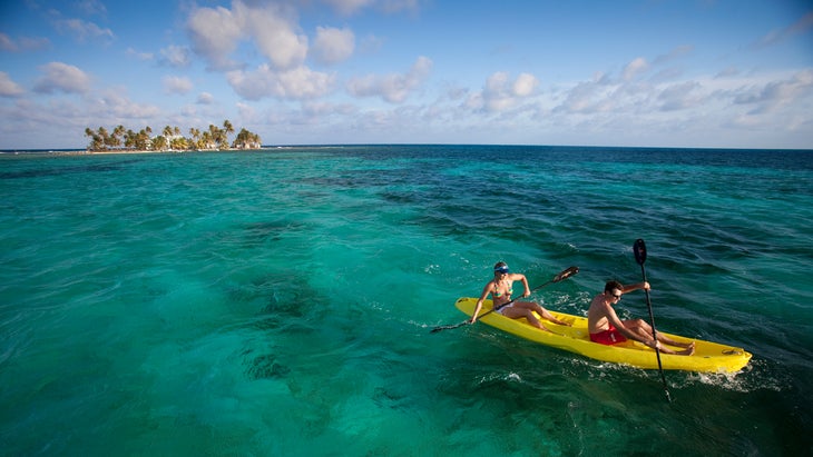 A couple paddling a yellow kayak in 80 degree, vivid blue, Caribbean water with a small tropical Island in the background.