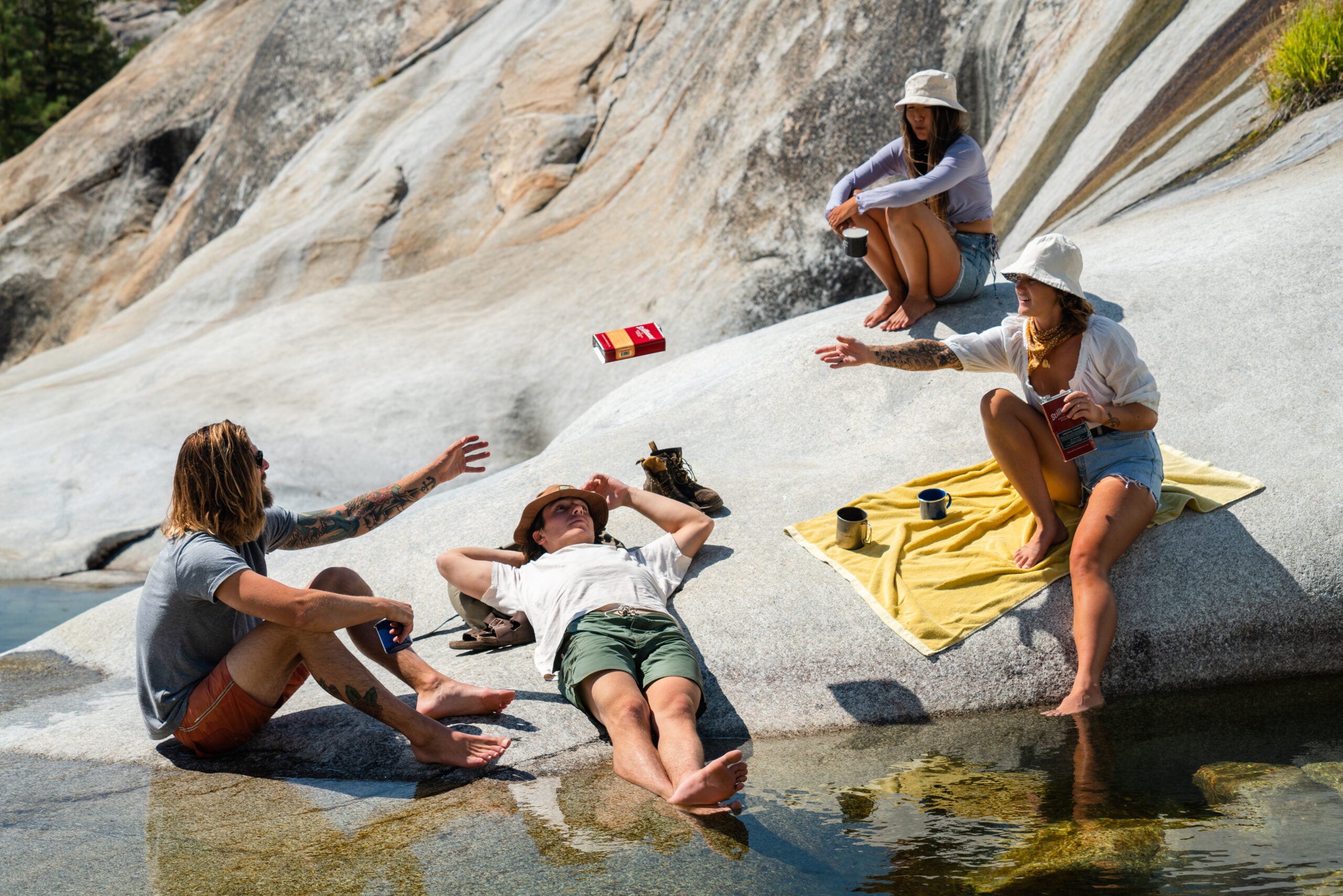 Several people enjoy a summer day on rocks by a river. One tosses a can of Stillhouse whiskey to another.