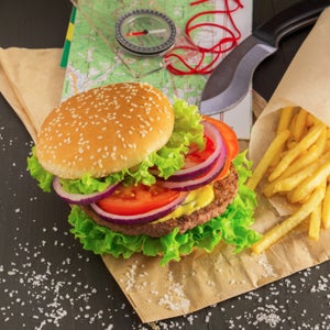 A juicy hamburger and fries sit on a table with a map, compass, and pocket knife.