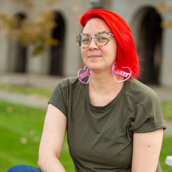 Frankie de la Cretaz is sitting outdoors. They have bright red hair and are wearing jeans, a green t-shirt, and pink earrings that say
