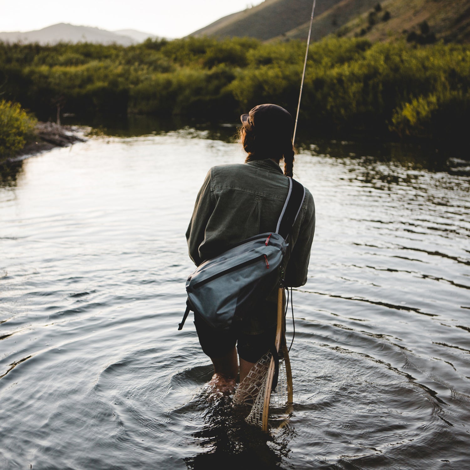 A woman fly fishing a small stream, her back is to us and she is carrying a small grey sling bag. It is sunset and there is golden light reflecting on the water.