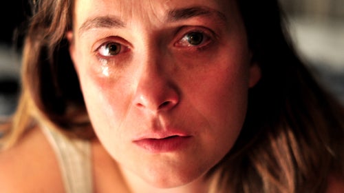 A white woman with blonde hair looks directly at the camera, crying quietly. She is wearing a white tank top.