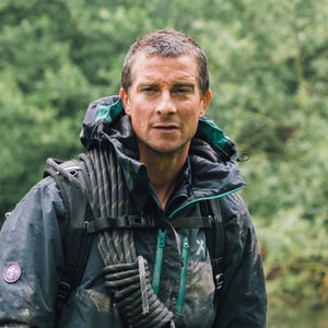 Bear Grylls stands in the forest wearing a dirty rain jacket, backpack, and climbing rope. He is squinting and not smiling.