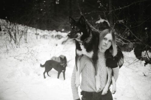 A black and white photo of a woman standing in a snowy backyard. She is carrying one dog on her shoulders, while another plays in the background.