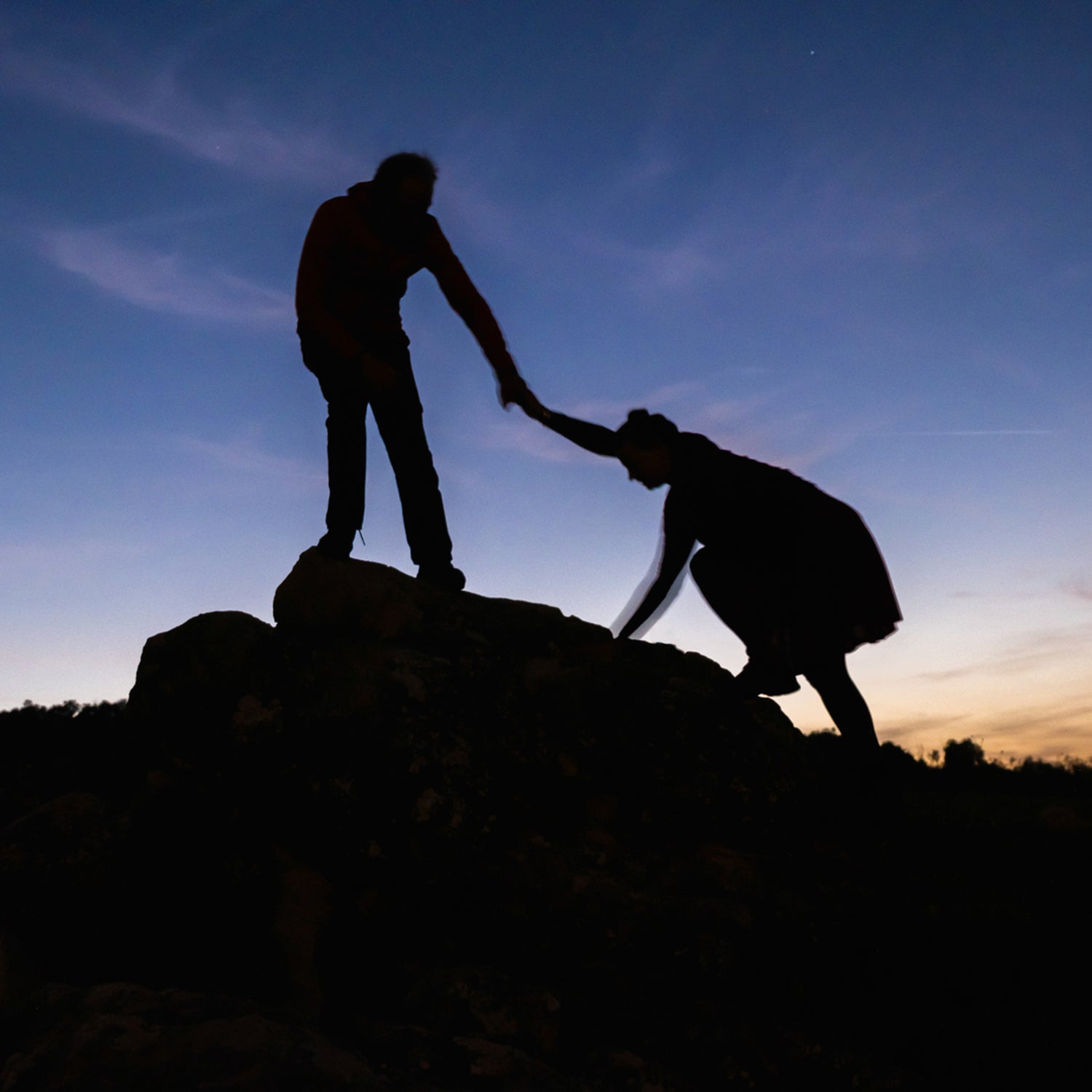 A silhouetted person helps another onto a rock