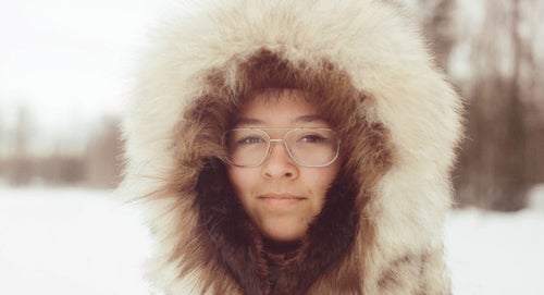 A woman wearing silver glasses and a furry hood sits in the snow.