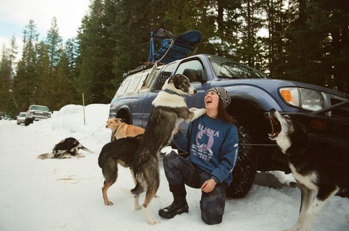 A woman kneeling in front of her vehicle laughing while surrounded by playful dogs