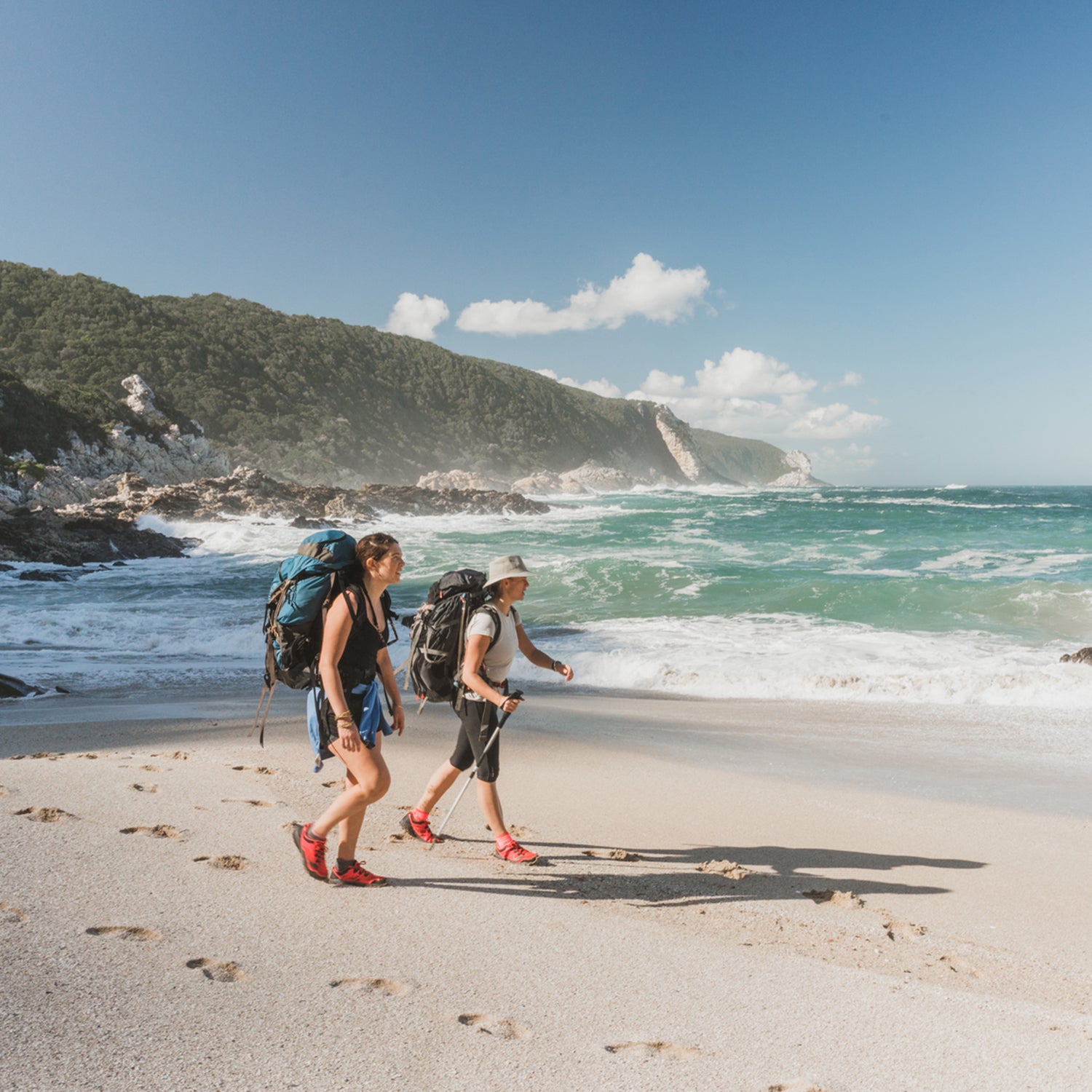 Two hikers carrying backpacks and a walking stick hiking across a scenic beach with waves crashing whilst on a coastal trail