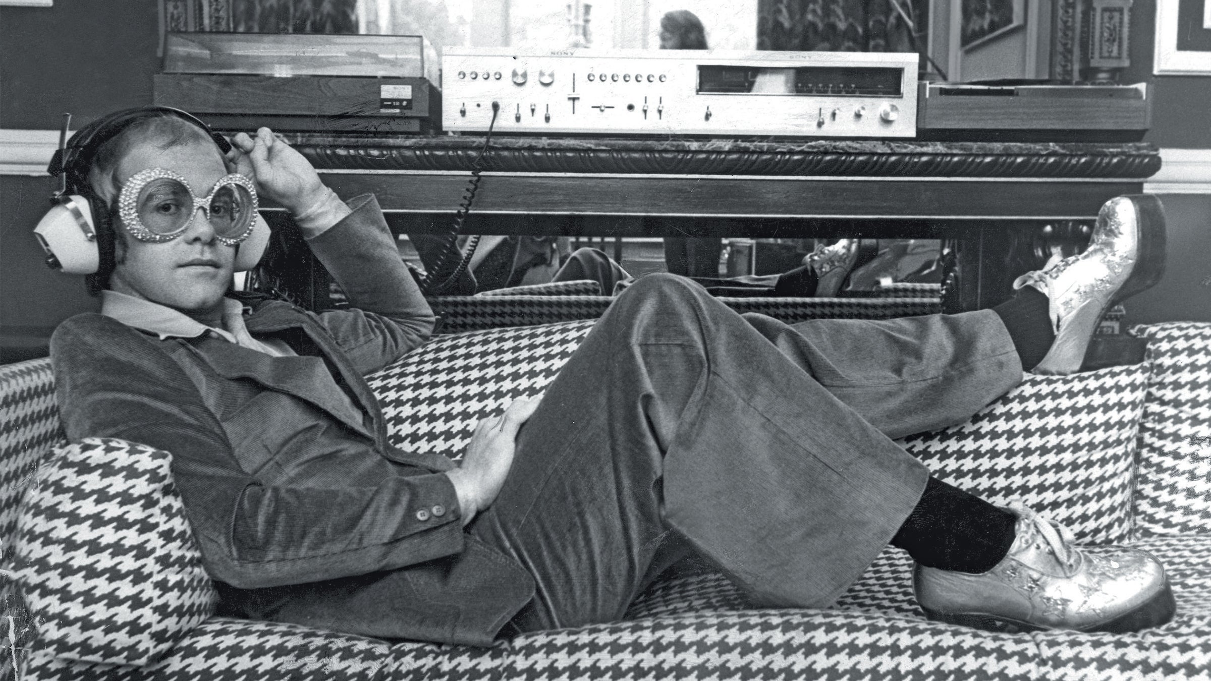 archival photo of Elton John, wearing sunglasses, listening to music on Sony hi-fi equipment