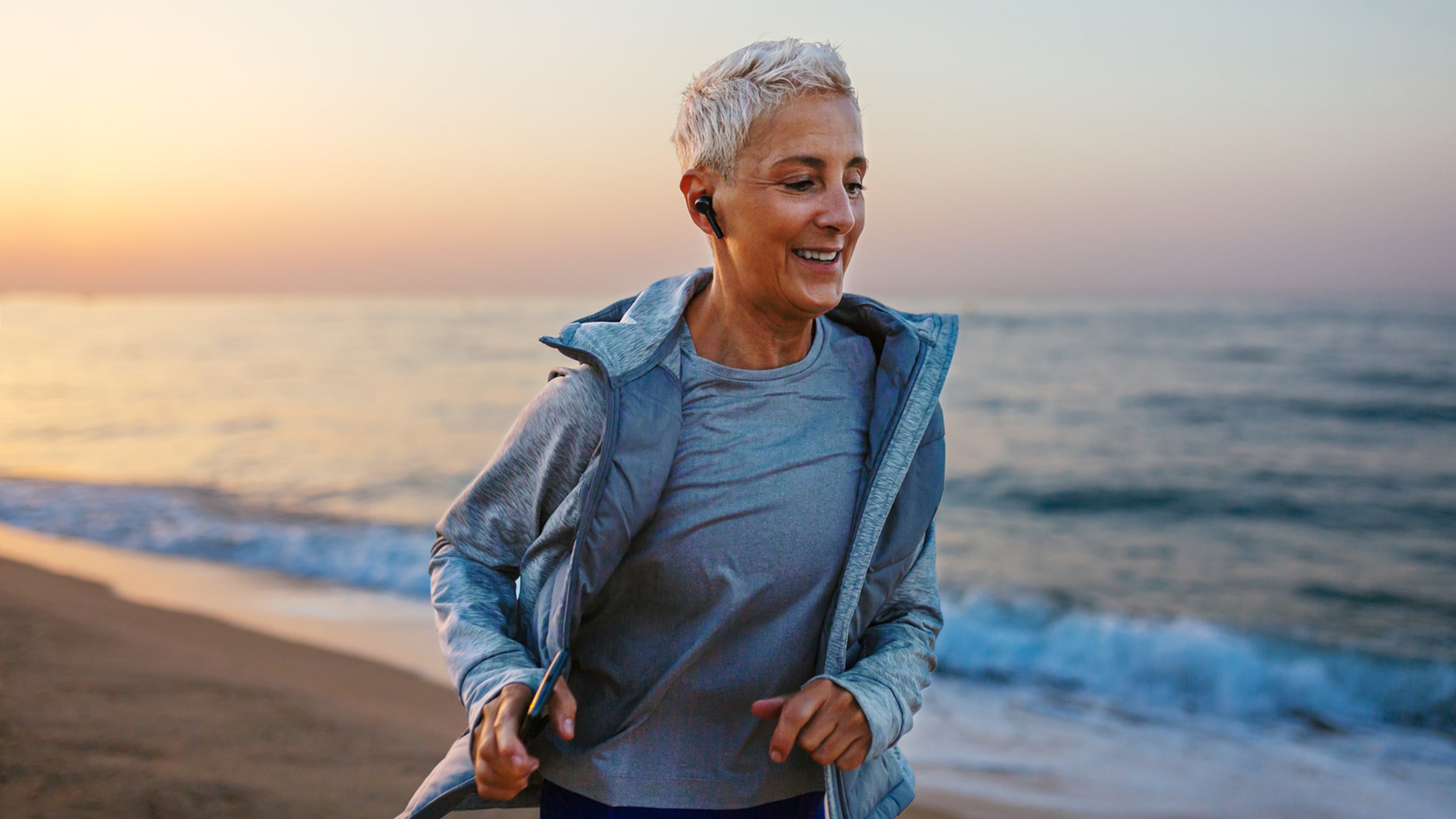 mature runner smiling and jogging on shore near waving sea at sunset