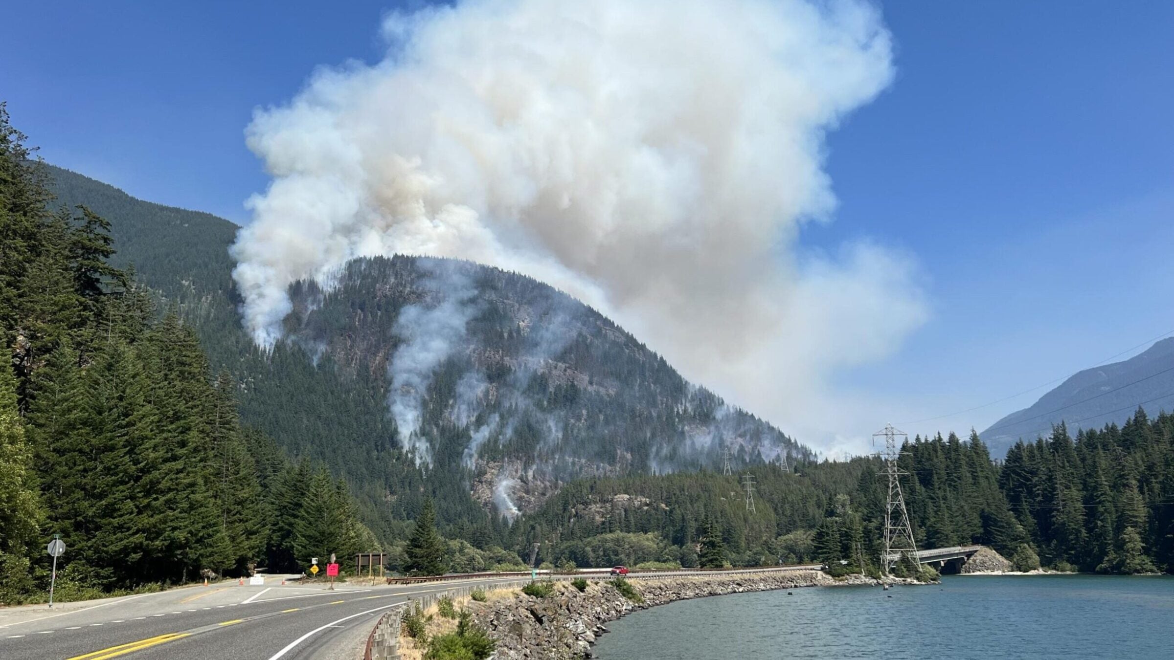 Wildfire smoke rises from a hillside next to a road running along a lake