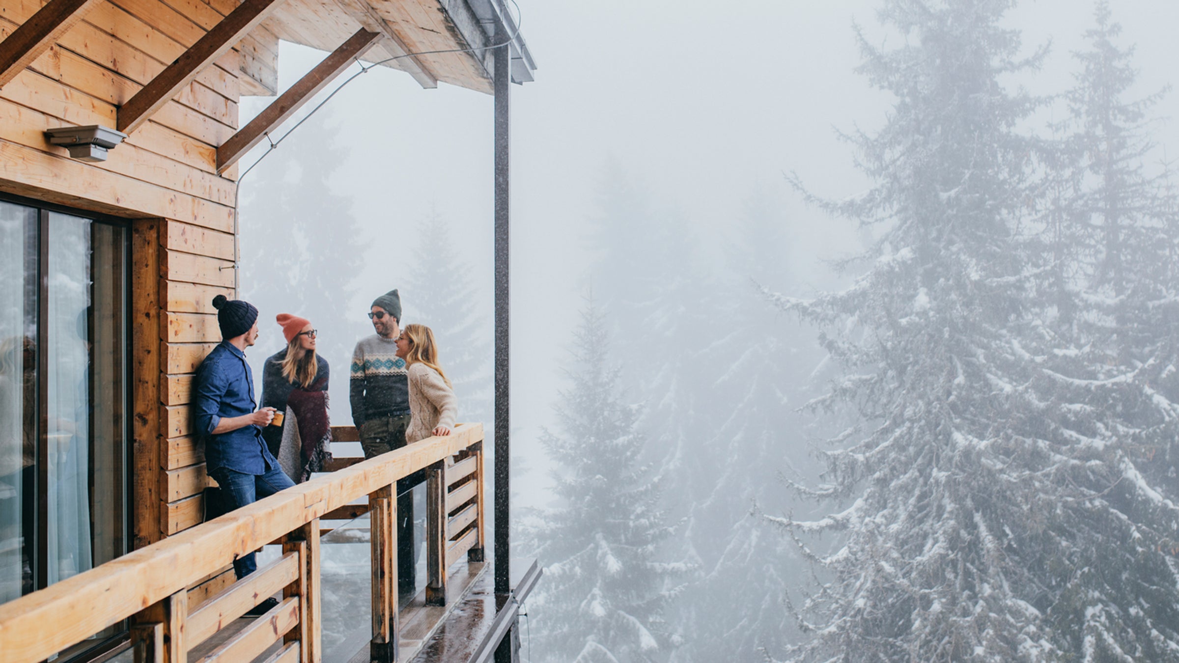 Group of men and women in winter outfits spending time on log cabin balcony in mountain.