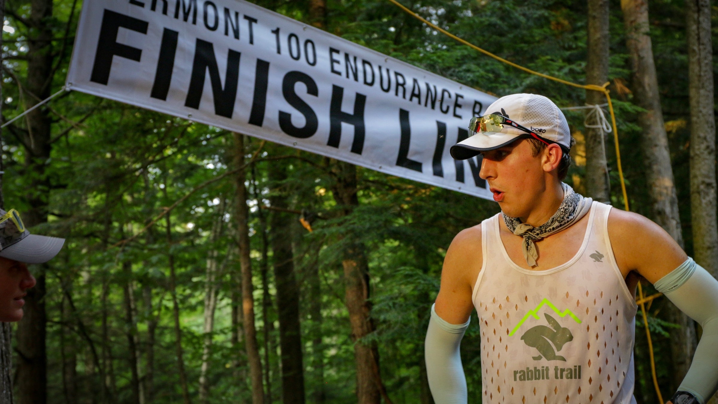 A man in compression sleeves, a tank top, and a baseball cap exhales at the finish line of the Vermont 100 endurance race