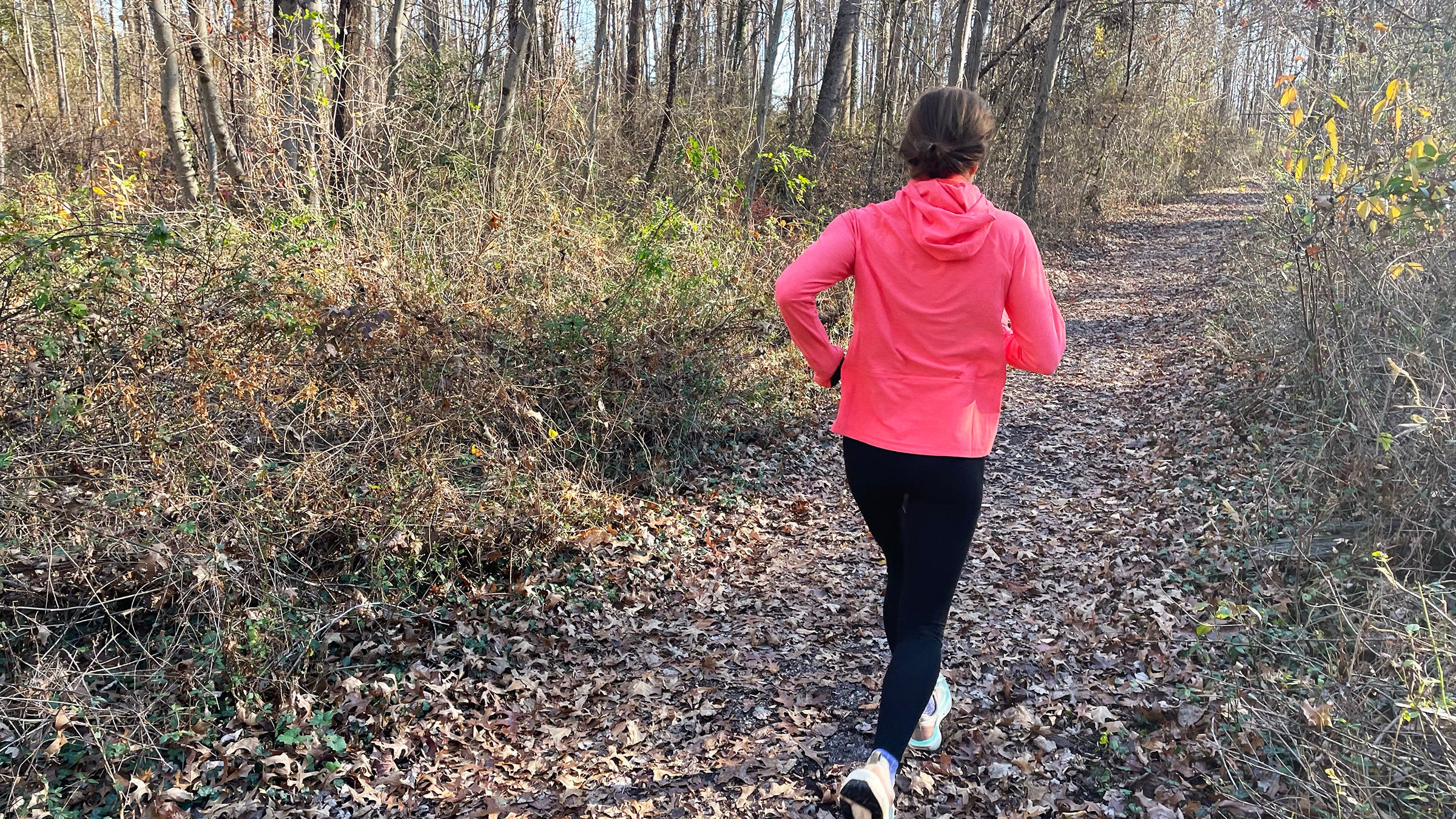 A runner moves along a leaf-covered forest trail wearing a bright pink running midlayer, captured from behind on a wooded path and photographed by author Lisa Jhung.