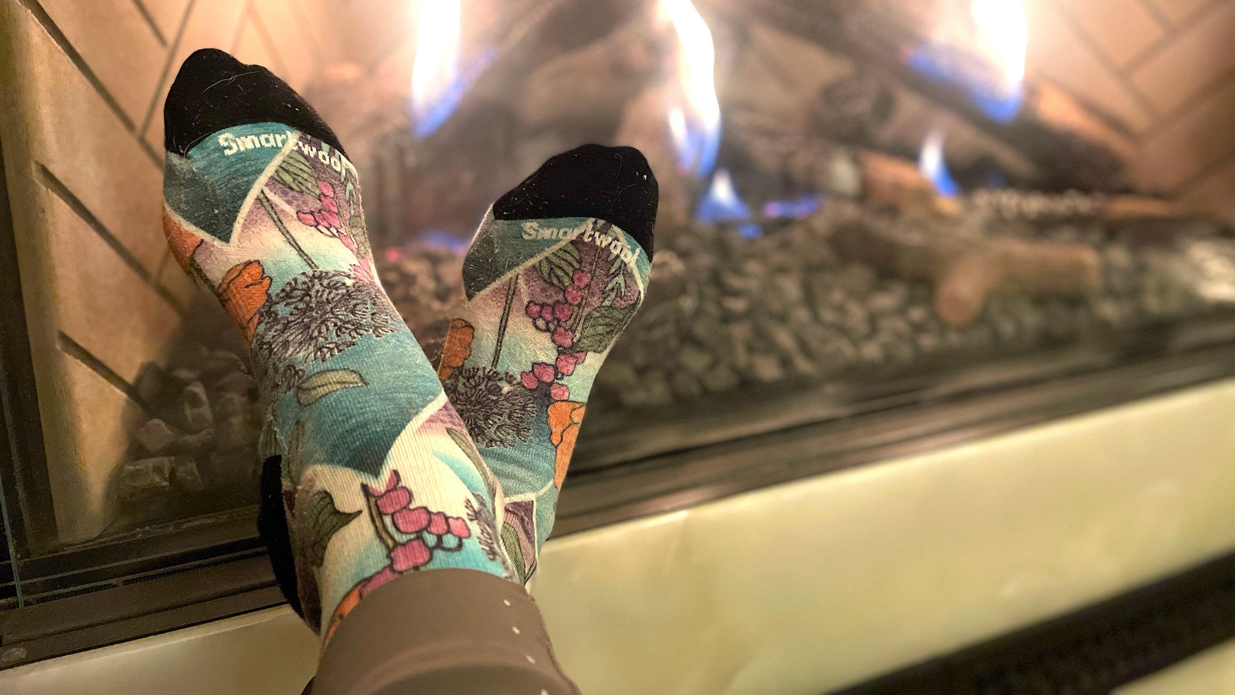 A close-up of colorful, botanical-patterned Smartwool socks being warmed in front of a glowing indoor fireplace.
