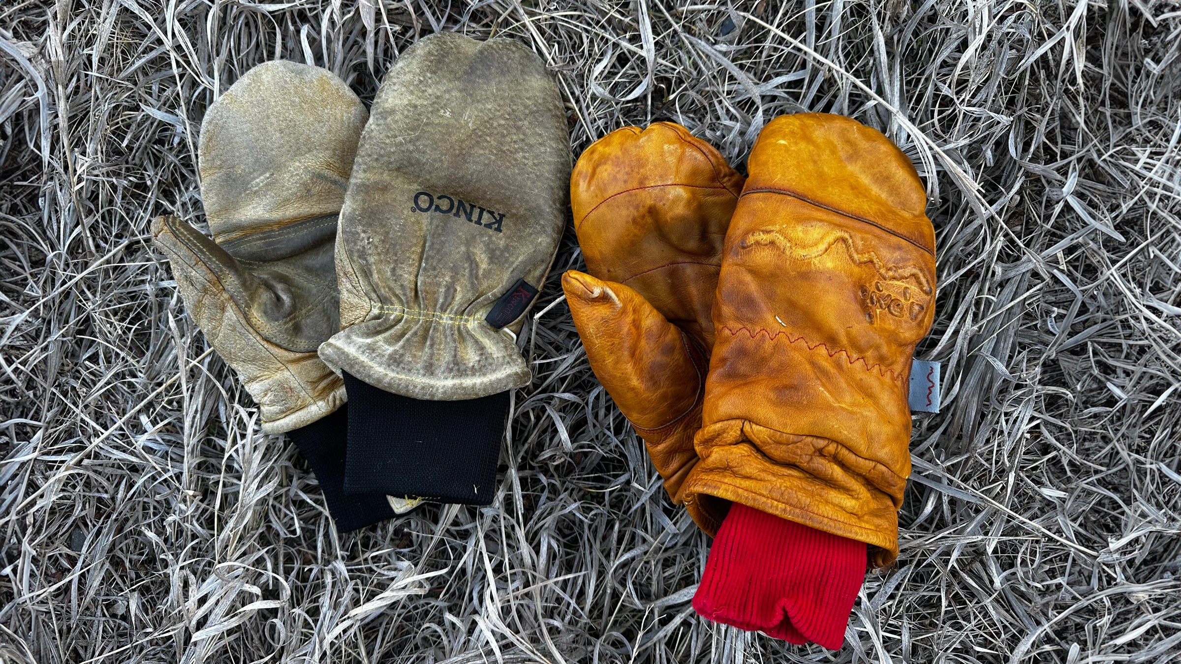 Close-up of two pairs of well-worn leather mittens resting on dry grass: the tan Give’r Frontier mitts with red knit cuffs on the right and the lighter Kinco Grain & Suede mitts with black cuffs on the left, showing natural creases and patina from heavy winter use.