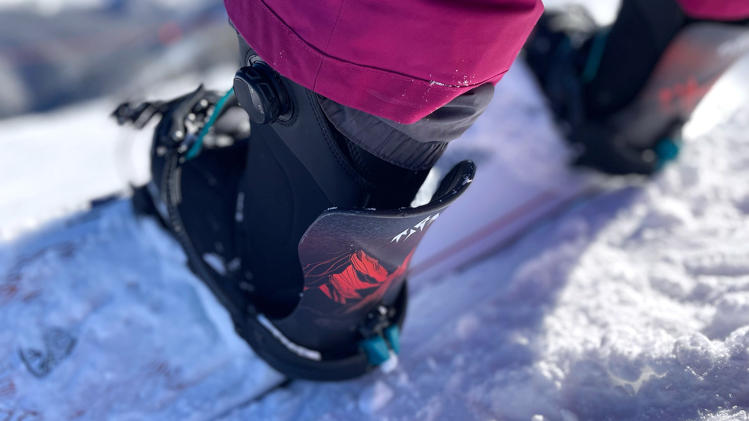 Close-up of a snowboard boot locked into the Jones Mercury FASE Snowboard Binding, showing the highback and dial-style FASE system set against a snowy mountain backdrop.